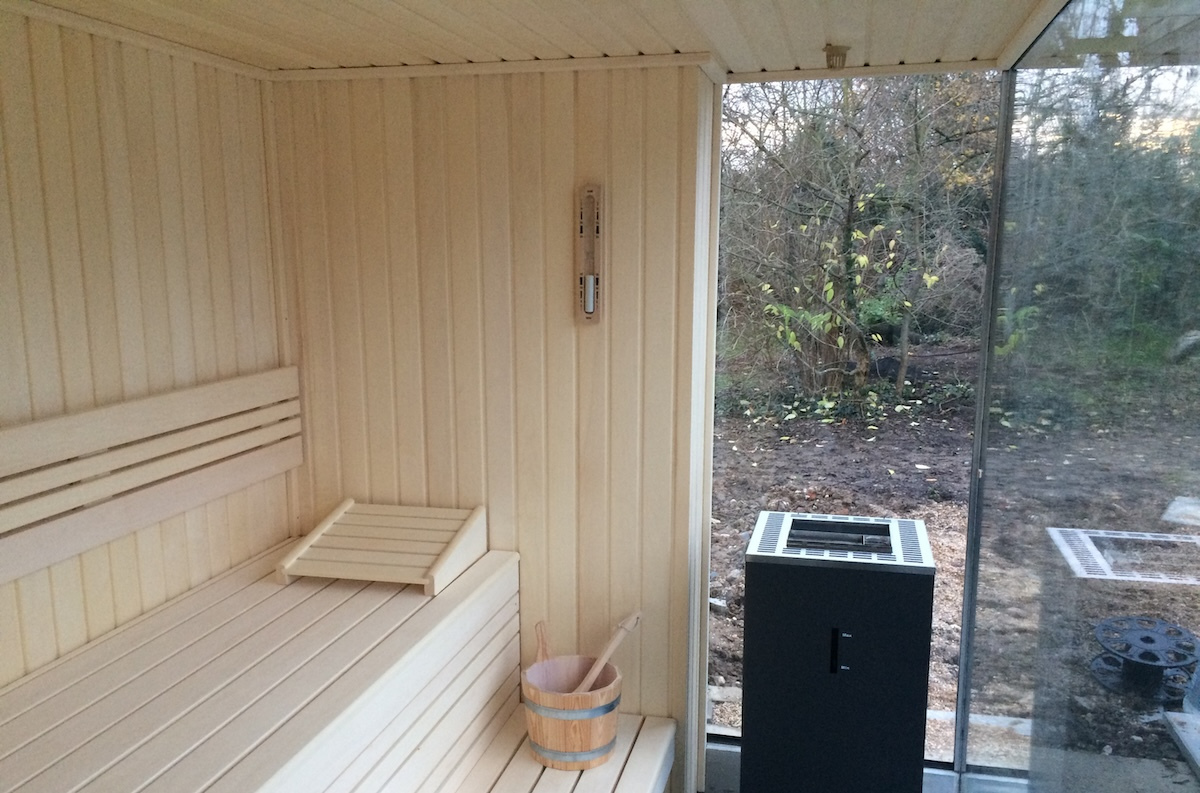 Sauna mit hellem Espenholz ausgekleidet, eingebaut in eine Gartenanlage mit freier Sicht in den Garten. Seitlich freistehender Saunaofen.